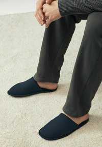 Des pantoufles en tissu bleu marine avec une texture lisse et un bout arrondi, portées par une personne assise sur un tapis de couleur claire, associées à un pantalon foncé.