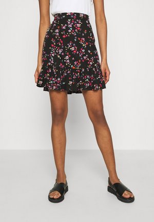 Kvinde iført sort blomster mini nederdel med flæsekant, hvid top og sorte sandaler står mod en ensfarvet baggrund.