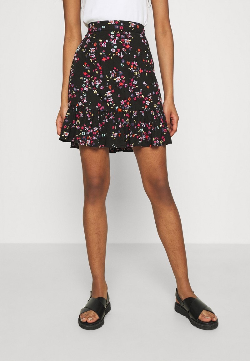 Femme portant une mini-jupe noire à motif floral avec un ourlet volanté, un haut blanc et des sandales noires, debout devant un fond uni.
