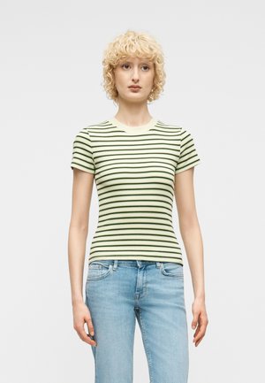 Νεαρό άτομο με κοντά, σγουρά ξανθά μαλλιά, που φοράει κοντομάνικη μπλούζα με πράσινες ανοιχτές και μαύρες ρίγες και ανοιχτόχρωμα μπλε τζιν, όρθιο.