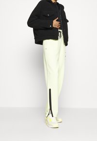 Black fleece jacket with two front pockets, paired with light yellow pants featuring side slits and black accents, worn with white sneakers.