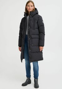 Lang svart pufferjakke med hette, med frontglidelås, to sidelommer og et merke på ermet. Brukt over en grå topp.