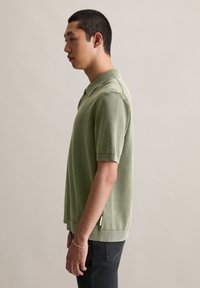 Mężczyzna w jasnoprzeładowej koszulce polo z krótkim rękawem i czarnych spodniach, stojący w profilu na jednolitym beżowym tle.