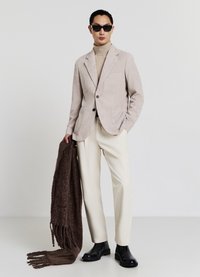 Hellbeiger Cordblazer, cremefarbener Rollkragenpullover, weiße Hose, brauner Schal mit Fransen, schwarze Stiefeletten und dunkle Sonnenbrille.