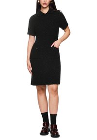 Schwarzes, kurzärmeliges, gestricktes Kleid mit Kragen, strukturierter Oberfläche, zwei vorderen Taschen und tailliertem Schnitt. Kombiniert mit schwarzen Socken und dunklen Schuhen.