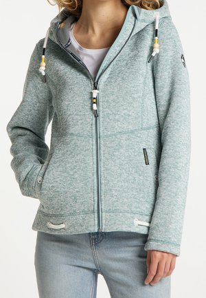 Femme portant un sweat à capuche zippé vert clair avec cordons et un jean bleu, debout avec une main dans la poche et l'autre détendue.