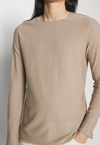 Haut beige à manches longues en tissu doux et texturé. Présente un col rond et des fentes latérales pour le confort et la liberté de mouvement.