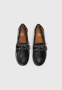 Zapatos de barco de cuero tejido negro con ojales y cordones dorados, que presentan una puntera suave y un cuerpo texturizado. Plantilla marrón visible en el interior.