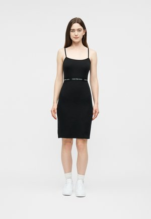 Nő áll, fekete, testhez simuló, ujjatlan ruha vékony pántokkal, fehér sportcipőben, egyszerű világos háttér előtt.