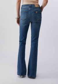 Femme portant un jean évasé bleu moyen et un haut sans manches côtelé beige, debout les mains détendues le long du corps, sur fond blanc.