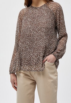 Frau trägt eine braune Langarmbluse mit weißen Punkten und beige Hosen, eine Hand in der Tasche, steht vor einem schlichten Hintergrund.