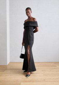 Robe noire décolletée à épaules nues avec un corsage ajusté et une fente haute, présentant un ourlet évasé. Accessoirisée avec des escarpins noirs à bouts pointus et un petit sac.