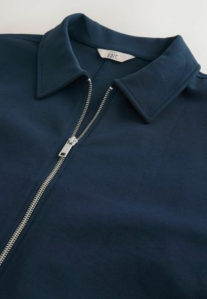 Tummansininen vetoketjullinen takki, jossa on terävä kaulus, valmistettu pehmeästä kankaasta. Keskellä hopeinen vetoketju. Yksinkertainen design.