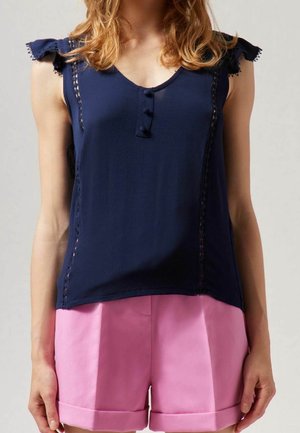 Frau trägt ein ärmelloses marineblaues Oberteil mit Knopfdetails und gerafften Schultern, kombiniert mit pinkfarbenen, umgeschlagenen Shorts.