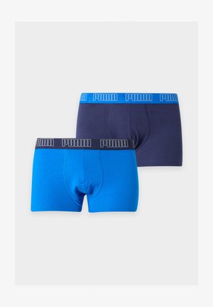 Homme mince à la peau foncée, debout pieds nus devant un fond uni, portant un boxer bleu marine avec une ceinture Puma bleue.
