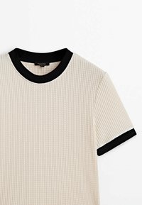 Chemise beige à manches courtes en maille gaufrée avec col rond noir et poignets de manches, présentée à plat sur un fond blanc.