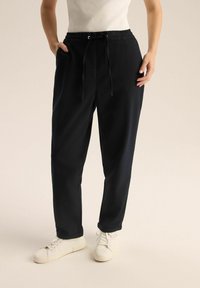 Granatowe spodnie dresowe z elastycznym pasem, detalem w postaci sznurka oraz bocznymi kieszeniami, w połączeniu z białymi sneakersami. Gładka tekstura materiału.