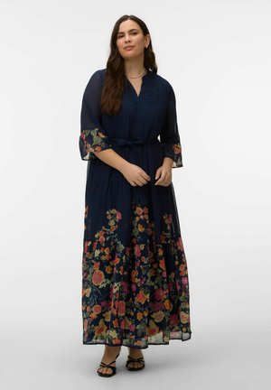 Femeie purtând o rochie lungă albastru închis cu modele florale pe mâneci și fustă, stând cu mâinile liniștit împreunate, sandale fără toc vizibile.