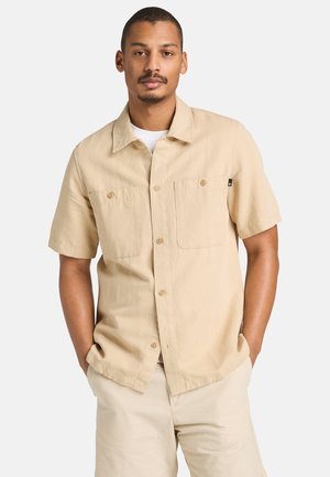 Uomo con camicia beige a maniche corte con due tasche sul petto, mani nelle tasche, in piedi contro uno sfondo chiaro e uniforme.