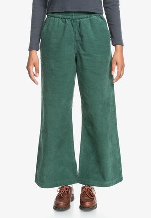 Personne portant un pantalon large en velours côtelé vert, des chaussures à lacets en cuir marron et un haut à manches longues foncé, debout face à l'avant.