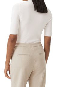Mujer con cabello negro lacio que lleva una blusa blanca de manga corta y pantalones beige a medida, vista desde atrás sobre un fondo blanco.