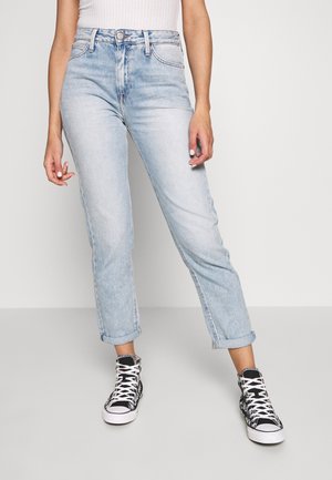 Femme portant un jean bleu clair roulé, un haut sans manches blanc côtelé et des baskets montantes noires avec des lacets blancs, debout devant un fond uni.