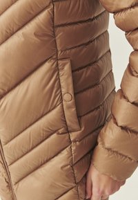 Doudoune marron avec un motif matelassé en chevrons, dotée d'une poche à bouton et d'une texture lisse. Gros plan sur la vue latérale.