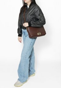 Borsa a spalla in pelle marrone con una forma strutturata, cuciture accentuate e una chiusura frontale. Indossata con una giacca bomber nera e jeans azzurri chiari.