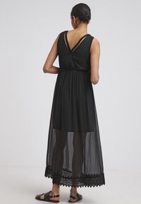 Svart klänning med figursydd överdel och ett flödande genomskinligt lager. Inkluderar en mönstrad spetskant och dubbla axelremmar bak. Lättviktsmaterial.