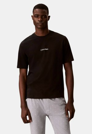 Hombre vistiendo una camiseta de manga corta negra de Calvin Klein y pantalones gris claro con patrones impresos sutiles, posando contra un fondo liso.