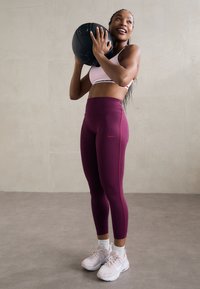 Sportiška moteris rožinės sportinės liemenėlės ir violetinių leggingų, laikanti juodą medicininį kamuolį, stovi ant pilko grindų prieš neutralų sienos foną.