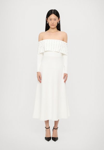Off-the-shoulder white dress with a textured, scalloped neckline and long sleeves, featuring a fitted bodice and flared skirt. Black heels complete the look.