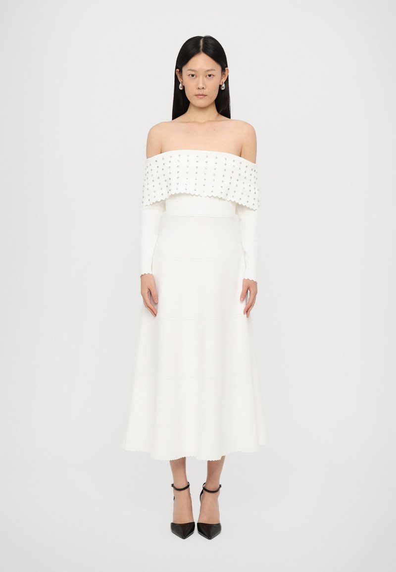 Off-shoulder witte jurk met een gestructureerde, gegolfde halslijn en lange mouwen, met een getailleerde bovenkant en een uitlopende rok. Zwarte hakken maken de look compleet.