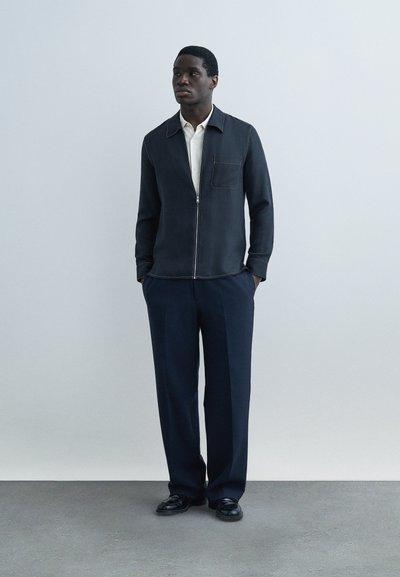 Άνδρας που στέκεται με τα χέρια στις τσέπες, φοράει σκούρο μπουφάν με φερμουάρ πάνω από λευκό πουκάμισο, άνετο σκούρο παντελόνι και μαύρα παπούτσια, μπροστά σε απλό φόντο.