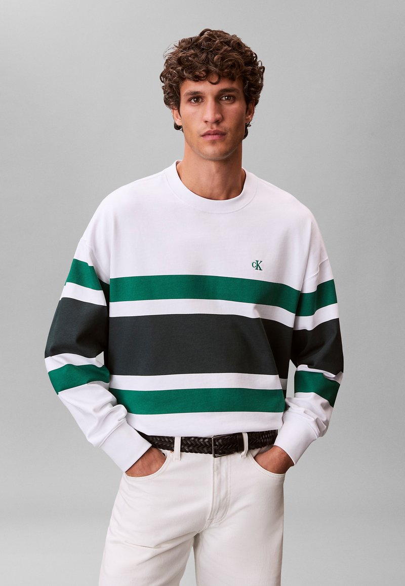 Jeune homme aux cheveux bouclés portant un pantalon blanc et un sweat-shirt blanc à rayures horizontales vertes et noires, les mains dans les poches.