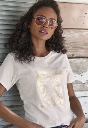 Light beige t-shirt featuring a gold foil botanical design on the front. Short sleeves and a round neckline, displayed against a rustic background.