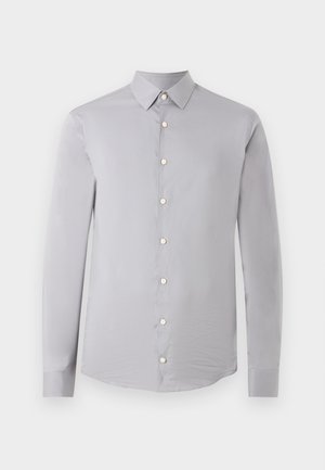 Uomo in piedi con la mano sinistra in tasca, indossa una camicia grigio chiaro con bottoni, pantaloni neri e scarpe eleganti nere, su sfondo semplice.