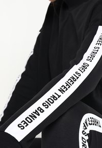 Sweatshirt noir avec des rayures blanches accentuées le long des manches et du texte. Le matériau est doux, avec une coupe décontractée et un design à col haut.