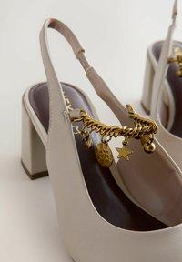 Escarpins beige à bride arrière avec talons blocs, semelles intérieures marron foncé et chaîne dorée décorative avec plusieurs breloques sur la lanière avant.