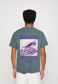 Grau-blaues Baumwoll-T-Shirt mit einem großen, bunten Hokusai-Druck auf dem Rücken, der einen Berg, Kraniche und stilisierte Wolken darstellt.