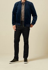 Granatowa bomberka z zamszu z prążkowanymi mankietami, w zestawieniu z brązową, wzorzystą koszulą, ciemnymi spodniami i czarnymi sneakersami. Prosty design, gładka faktura.