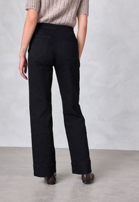 Osoba ubrana w czarne spodnie o prostych nogawkach i brązowe buty, stojąca na szarej podłodze na tle jasnoszarej ściany, widziana od tyłu.