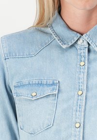 Camisa de denim azul claro con cuello puntiagudo, bolsillo frontal y botones a presión decorativos en tono dorado. Presenta una textura suave y un diseño clásico.