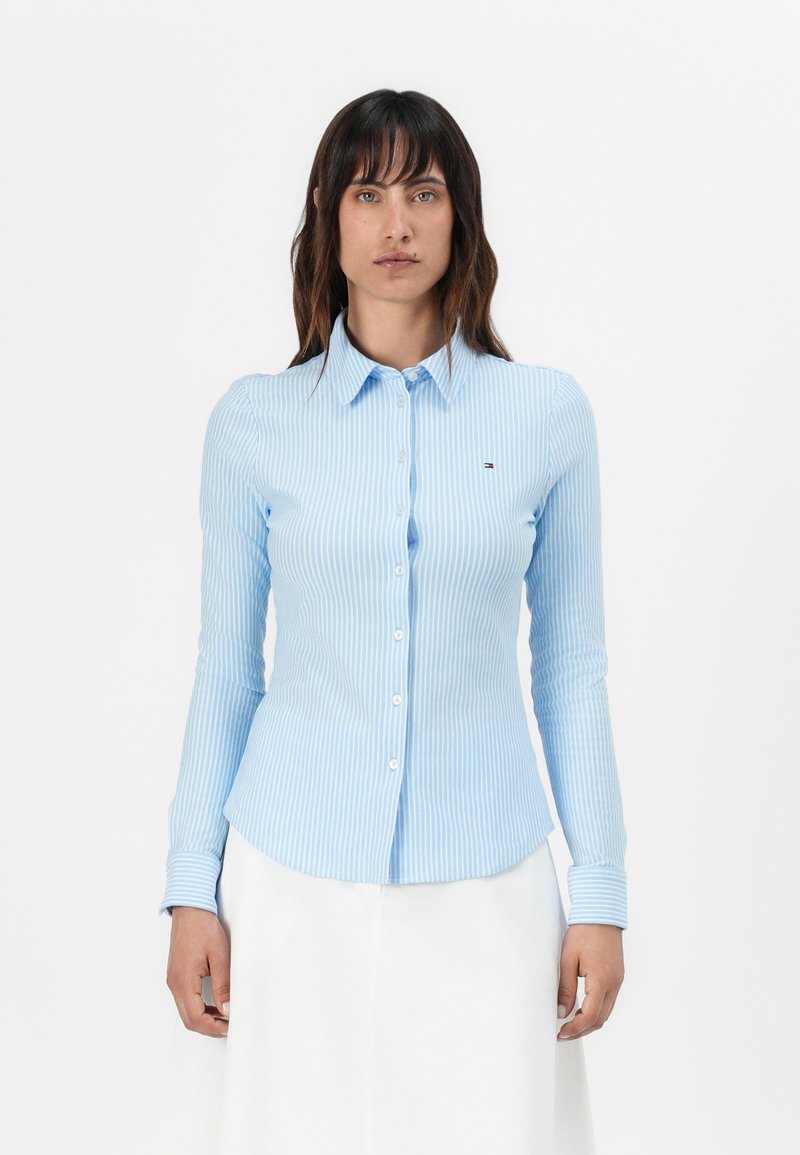 Camicia azzurra chiaro con maniche lunghe e bottoni, con righe verticali bianche. Presenta un colletto, un design aderente e un piccolo logo sul petto.