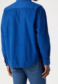 Blå långärmad skjorta med knäppkrage. Tillverkad av slät tyg, har sömnadsdetaljer och manschetter, med en rundad nederkant.