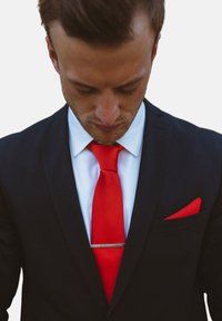 Chaqueta de traje azul marino con una camisa blanca, corbata roja y pañuelo de bolsillo rojo. Presenta un sujetacorbatas plateado. Se centra en la parte superior del cuerpo y los detalles de la vestimenta.