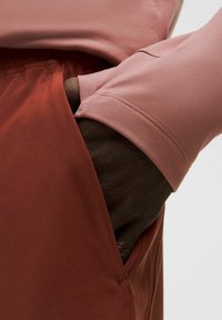 O mână cu pielea închisă la culoare odihnindu-se în interiorul buzunarului unor pantaloni de culoare ruginită, purtând un top cu mânecă lungă de un roz prăfuit, cu deschidere pentru degete.