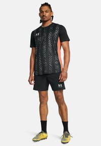Camiseta deportiva negra con textura de rayas verticales y panel lateral color coral, combinada con pantalones cortos negros y zapatillas de deporte amarillas.