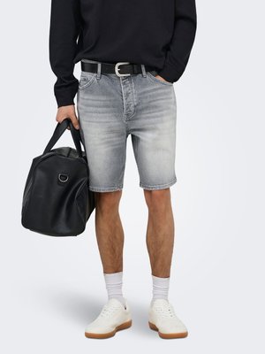 Uomo che indossa pantaloncini in denim grigio sbiadito, cintura nera, camicia nera a maniche lunghe, sneaker bianche e tiene in mano una borsa da viaggio in pelle nera.