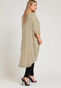 Beige oversized tunika med korta, vida ärmar; mjukt, slätt tyg; asymmetrisk nederkant; ihopparad med svarta figurnära jeans och svarta klackar.